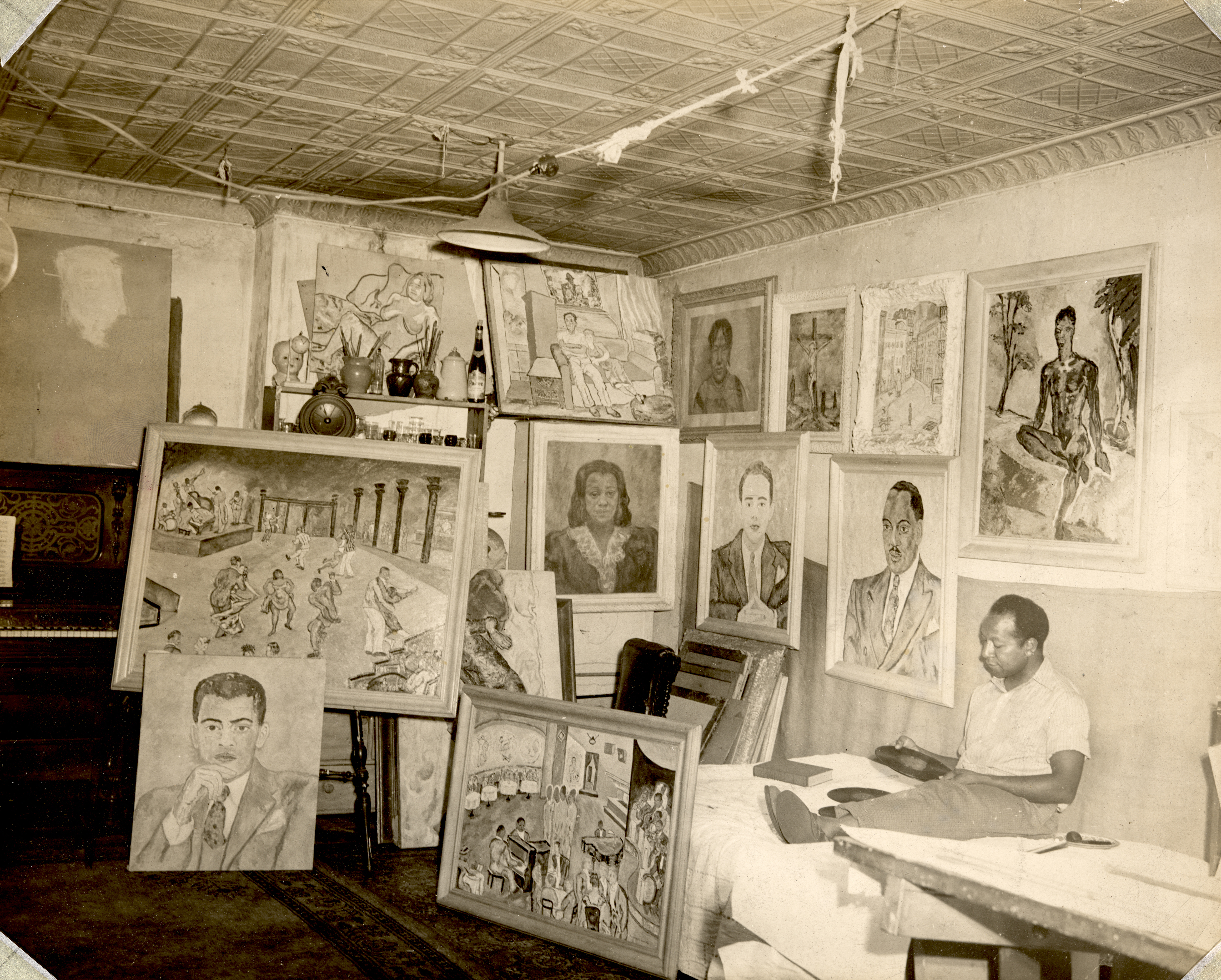 Photo of Beauford Delaney in his studio, 1940. Paintings behind him include portraits of musicians and a scene in the Savoy Ballroom. 