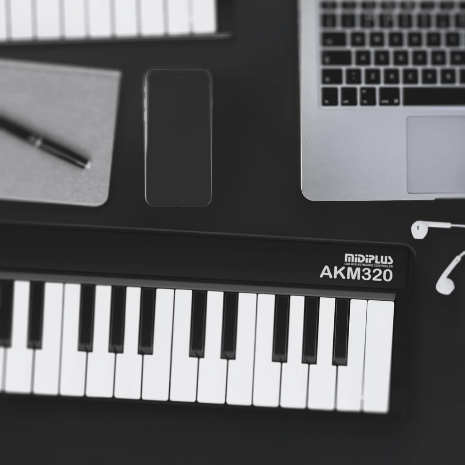 A MIDI keyboard on a desk with a phone, laptop, pen and paper, and headphones.