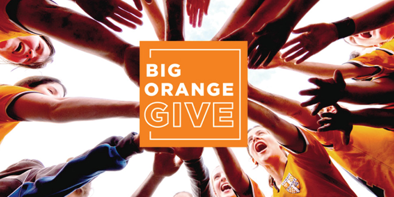 photo of a circle of students reaching into the center of the circle to touch hands (legend reads "Big Orange Give")